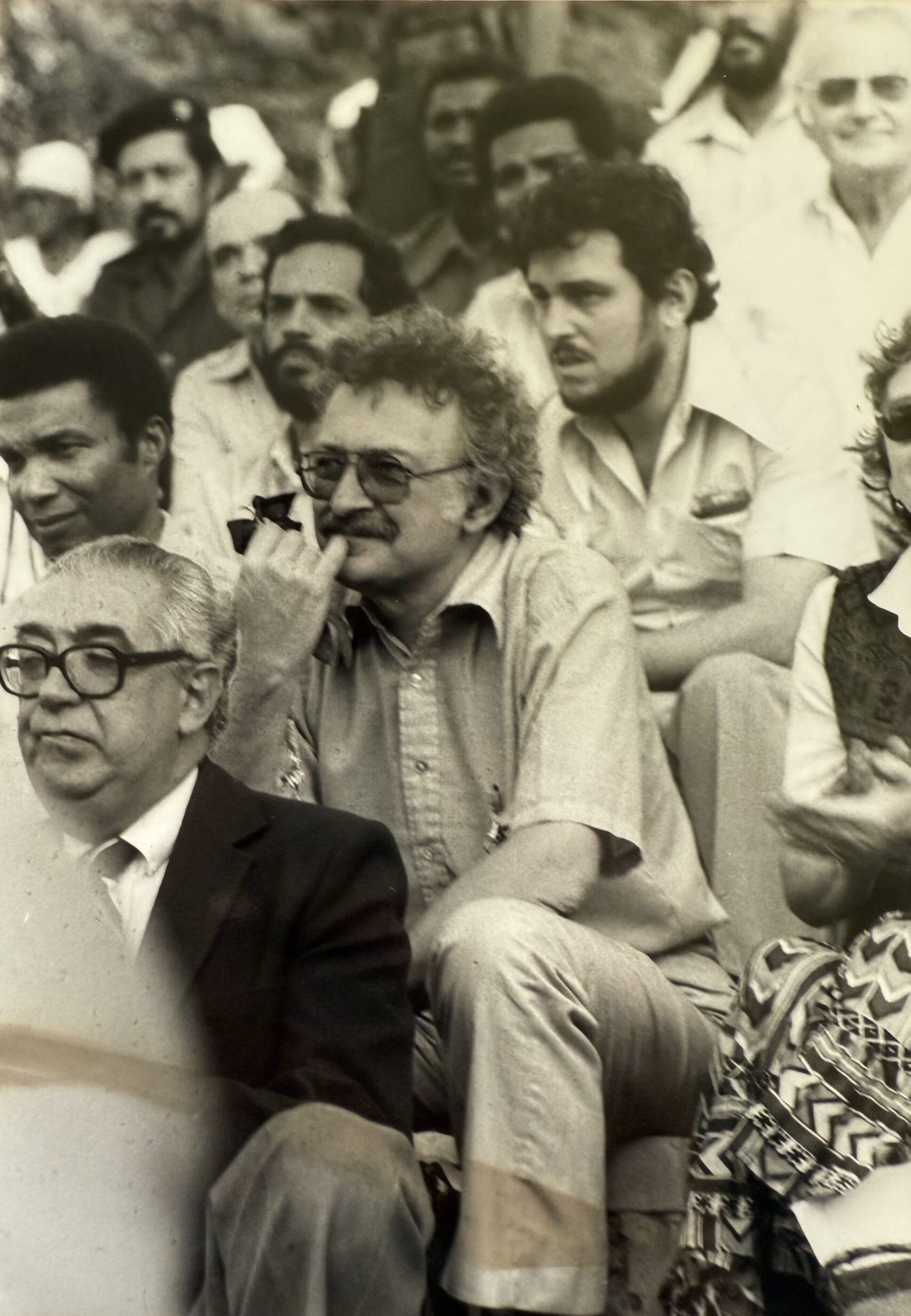 Black-and-white photograph of Immanuel Wallerstein seated in an outdoor audience, holding a pipe, among a crowd of seated listeners.